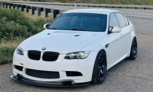 Białe BMW E90 M3 z czarnymi felgami i spoilerem, zaparkowane przy drodze z barierkami.