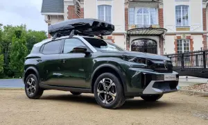 Nowy C3 Aircross w ciemnozielonym kolorze z czarnym boxem dachowym przed eleganckim budynkiem.