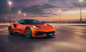Pomarańczowy Lotus Emira 420 Sport na torze wyścigowym o zachodzie słońca, z rozmazanymi światłami w tle.