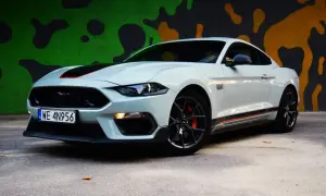 Biały Ford Mustang Mach 1 z czarnymi pasami i pomarańczowymi akcentami na tle kolorowego muralu.