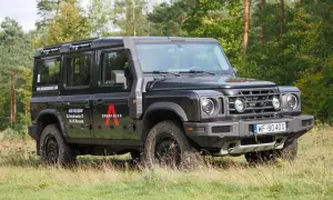 Czarny INEOS Grenadier z logo "A" na drzwiach, zaparkowany na trawie, z lasem w tle.