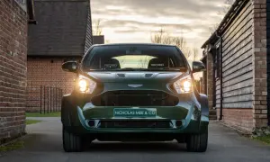 Aston Martin Cygnet w ciemnozielonym kolorze, z reflektorami świecącymi w stronę widza, zaparkowany na podjeździe między ceglanymi budynkami.