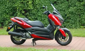 Czerwony skuter Yamaha XMAX 125, gotowy do odblokowania pełnej mocy, zaparkowany na tle zielonego żywopłotu.