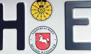 Fragment niemieckich tablic rejestracyjnych z literami H i E, naklejkami z regionu Hanower i datą przeglądu.