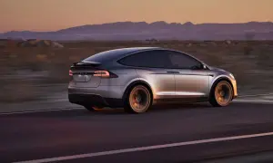 Srebrny Tesla X pędzi przez pustynię o zachodzie słońca, góry w tle.