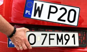 Ręka z bransoletką montuje tablicę rejestracyjną z napisem "O 7FM91" na czerwonym samochodzie. Obok widać inną tablicę "W P20".