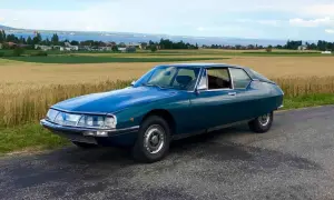 Piękny, turkusowy Citroen SM na tle złotego pola i jeziora. Klasyk motoryzacji w malowniczej scenerii.