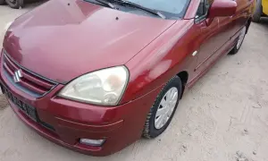 Czerwony Suzuki Liana stoi na piaszczystym podłożu. Widoczny przód auta z reflektorem i grillem.