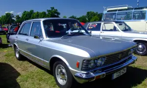 Srebrny Fiat 130 z lat 70. prezentuje się elegancko na trawie, obok innych klasyków motoryzacji.