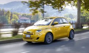 Nowy Fiat 500 w kolorze żółtym sunie po drodze, otoczony jesienną zielenią i drzewami.