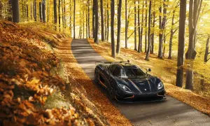 Mroczny Koenigsegg Agera RS z pomarańczowymi akcentami przemierza jesienną drogę wśród złotych liści i drzew.