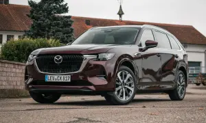 Elegancka, ciemnobordowa Mazda CX-80 stoi przed budynkiem z czerwoną dachówką.