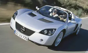 Srebrny Opel Speedster z parą na pokładzie mknie po krętej drodze, ciesząc się słońcem i wolnością.