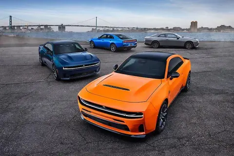Pomarańczowy i niebieski Dodge Charger, szary Dodge Challenger i niebieski Dodge Charger stoją nad wodą z mostem w tle.