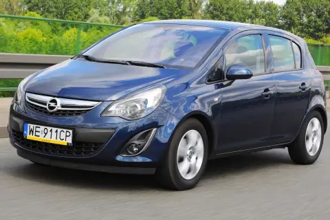 Granatowy Opel Corsa D jedzie po drodze, odbijając niebo i chmury.