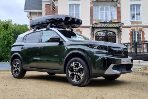 Nowy C3 Aircross w ciemnozielonym kolorze z czarnym boxem dachowym przed eleganckim budynkiem.