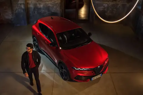 Elegancki mężczyzna w okularach obok lśniącego, czerwonego Alfa Romeo Tonale. Stylowy duet w industrialnym wnętrzu.