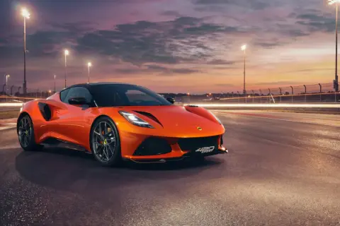 Pomarańczowy Lotus Emira 420 Sport na torze wyścigowym o zachodzie słońca, z rozmazanymi światłami w tle.