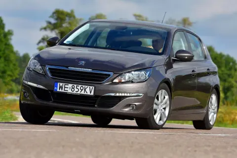 Ciemnoszary Peugeot 308 2015 z polskimi tablicami WE 859KV jedzie po drodze wśród zieleni.