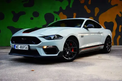 Biały Ford Mustang Mach 1 z czarnymi pasami i pomarańczowymi akcentami na tle kolorowego muralu.