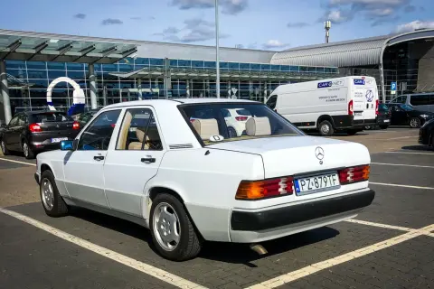 Biały Mercedes W190 z lat 80. parkuje przed nowoczesnym budynkiem lotniska. Klasyk w świetnym stanie.