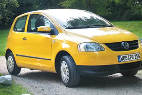 Żółty Volkswagen Fox zaparkowany na żwirowej drodze, otoczony zielenią.