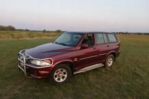 Ciemnoczerwony Daewoo Musso z orurowaniem z przodu stoi na trawiastym polu. W tle jezioro i księżyc.