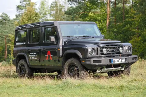Czarny INEOS Grenadier z logo "A" na drzwiach, zaparkowany na trawie, z lasem w tle.