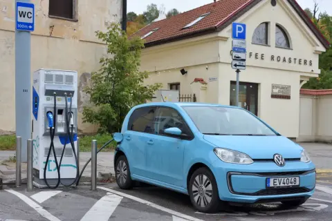 Niebieski Volkswagen Up ładuje się przy stacji ładowania. Obok znajduje się budynek z napisem "COFFEE ROASTERS".