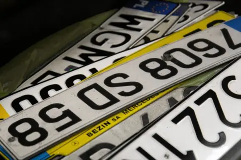 Stos tablic rejestracyjnych, w tym jedna z napisem "BEZIN SA MERCEDES". Widać fragmenty numerów, np. 8509L, 85DS, WGM.