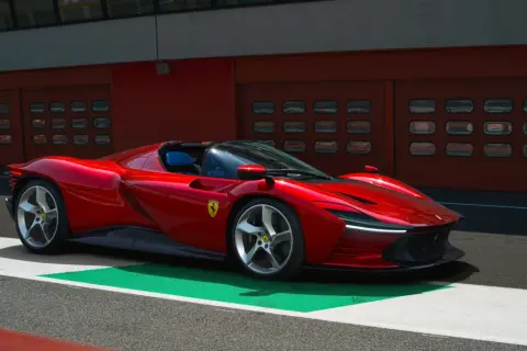 Czerwone Ferrari SP3 na torze wyścigowym, z niebieskimi siedzeniami i garażami w tle.