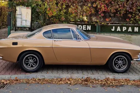 Klasyczny Volvo P1800 w kolorze złotym, zaparkowany na chodniku, z jesiennymi liśćmi wokół.