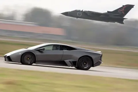 Szary Lamborghini Reventon pędzi po pasie startowym, gdy myśliwiec Tornado przelatuje nad nim.
