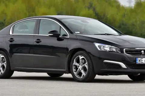 Elegancki, czarny Peugeot 508 z chromowanymi detalami i felgami stoi na tle zieleni.