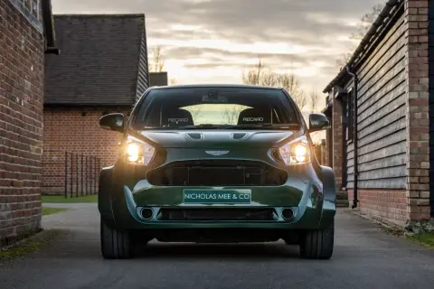 Aston Martin Cygnet w ciemnozielonym kolorze, z reflektorami świecącymi w stronę widza, zaparkowany na podjeździe między ceglanymi budynkami.