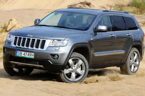 Szary Jeep Grand Cherokee 3.6 pokonuje piaszczysty teren, wzbijając tumany kurzu.