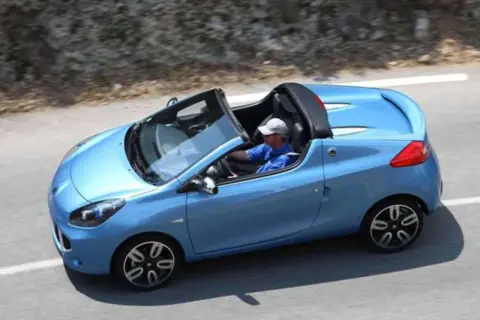Niebieski kabriolet Renault Wind z otwartym dachem, kierowca w czapce i niebieskiej koszulce jedzie po krętej drodze.