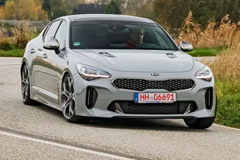 Szary Kia Stinger z kierowcą za kierownicą jedzie po krętej drodze, otoczony jesienną roślinnością.