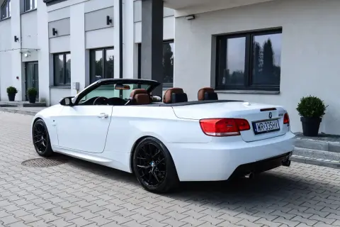 Białe BMW E93 cabrio z brązową skórzaną tapicerką i czarnymi felgami stoi na kostce brukowej przed nowoczesnym budynkiem.