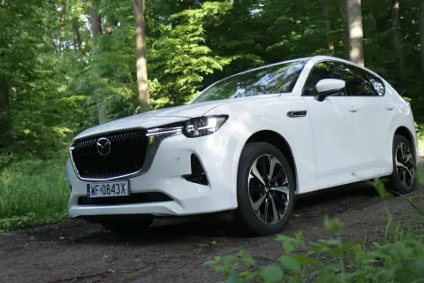 Biała Mazda CX-60 zaparkowana na leśnej ścieżce, otoczona zielenią drzew i traw.