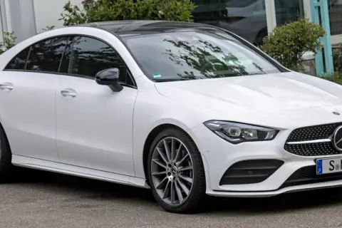 Biały Mercedes CLA 180 z czarnym dachem i przyciemnianymi szybami stoi na tle zieleni.