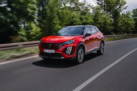 Czerwony Peugeot 2008 GT pędzi po drodze, otoczony zielenią drzew.