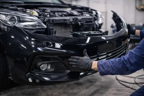 Mechanik w czarnej rękawiczce przykłada czarny zderzak do przodu samochodu.