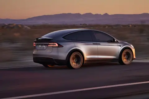 Srebrny Tesla X pędzi przez pustynię o zachodzie słońca, góry w tle.