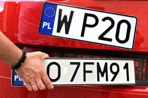 Ręka z bransoletką montuje tablicę rejestracyjną z napisem "O 7FM91" na czerwonym samochodzie. Obok widać inną tablicę "W P20".