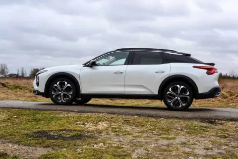 Biały Citroen C5X na tle pochmurnego nieba i wiejskiego krajobrazu. Elegancki design i nowoczesne felgi podkreślają jego wyjątkowość.