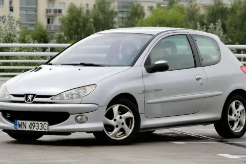 Srebrny Peugeot 206 Plus z napisem "Quiksilver" na błotniku, zaparkowany na mokrym parkingu.
