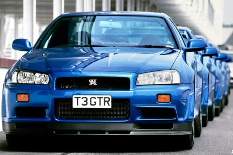 Błękitny Nissan Skyline GT-R R34 z tablicą "T3 GTR" ustawiony w rzędzie z innymi.