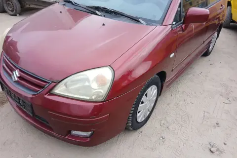 Czerwony Suzuki Liana stoi na piaszczystym podłożu. Widoczny przód auta z reflektorem i grillem.
