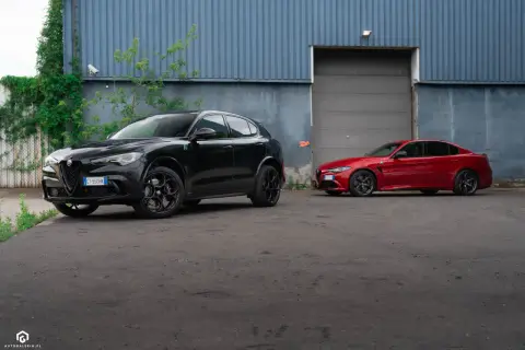 Dwa sportowe Alfa Romeo: czarny Stelvio Quadrifoglio i czerwona Giulia, stoją przed budynkiem z niebieską, falistą fasadą.